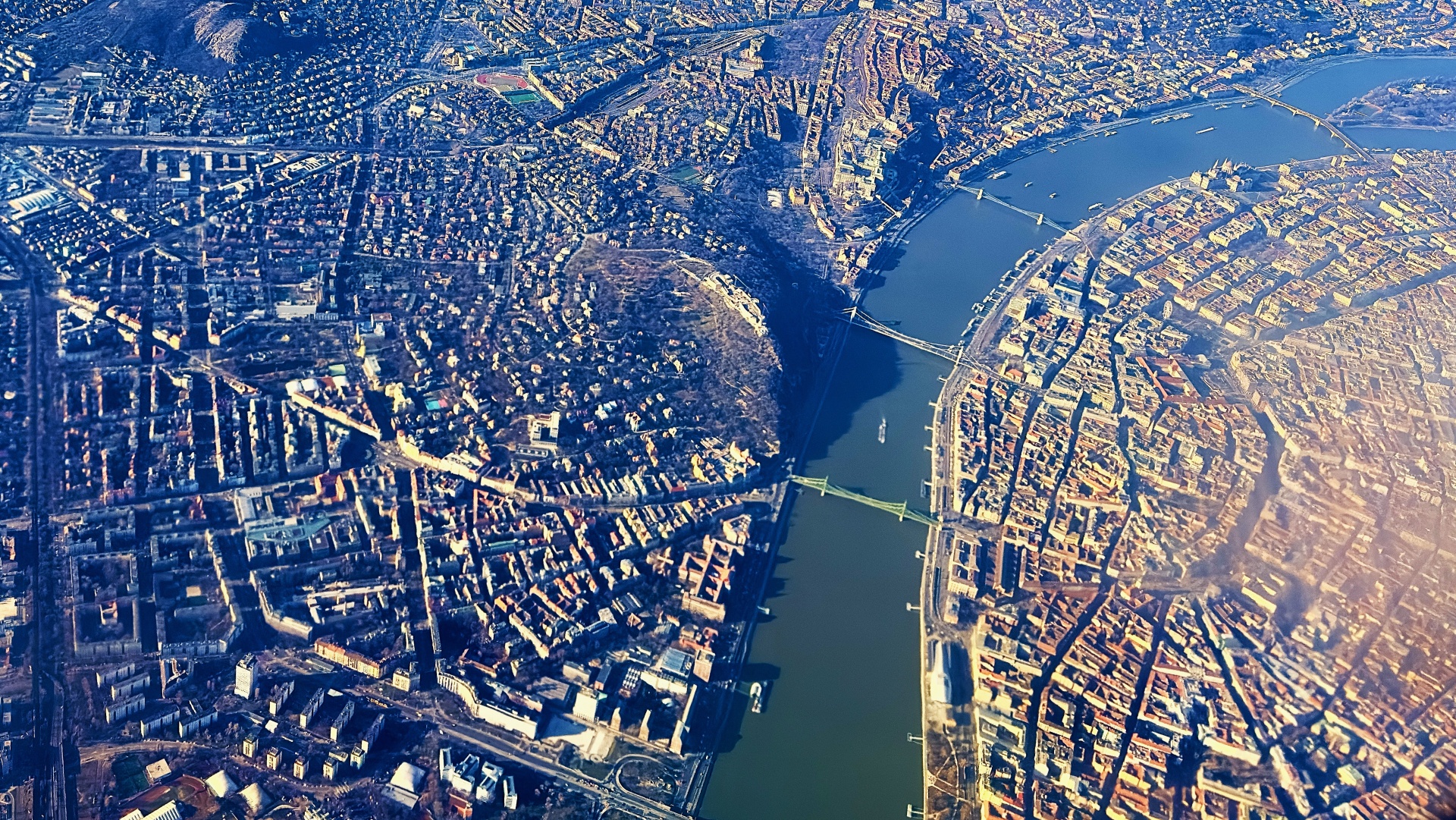 A wide river with two boats sailing, flanked by lush green trees and houses, with a bridge and hills in the background under a clear sky.