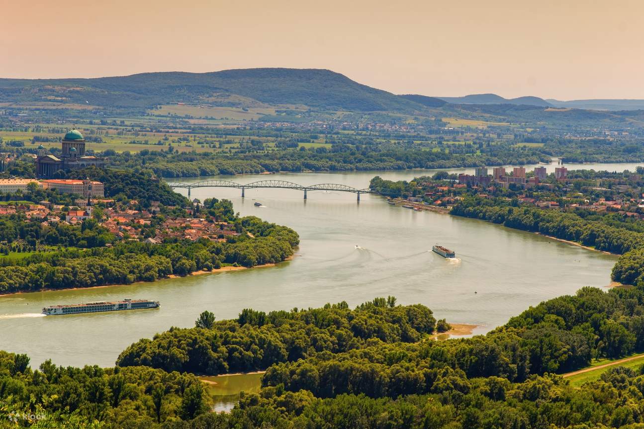 Aerial view of a river with two boats and a bridge, surrounded by green trees, a town with red-roofed buildings, and hills in the background under a clear sky.