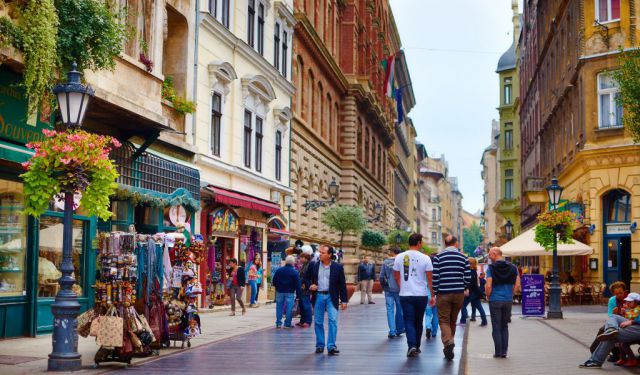 Crowd crossing a bridge decorated with colorful flags and a large Sziget Island of Freedom festival sign overhead under a clear blue sky.