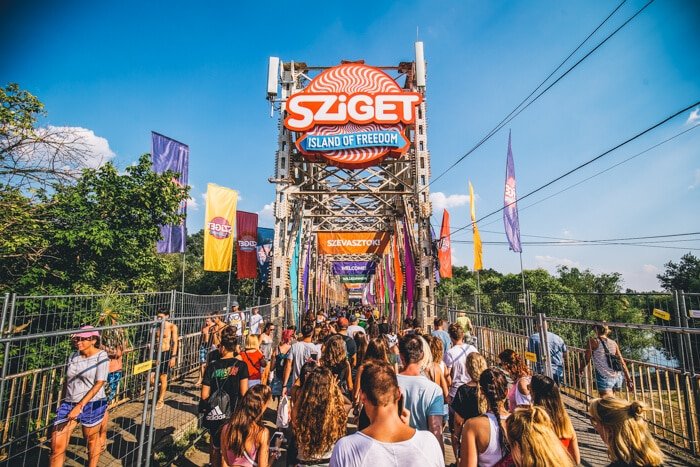 Crowd walking toward entrance of Sziget Festival on a bridge decorated with colorful flags under a blue sky.