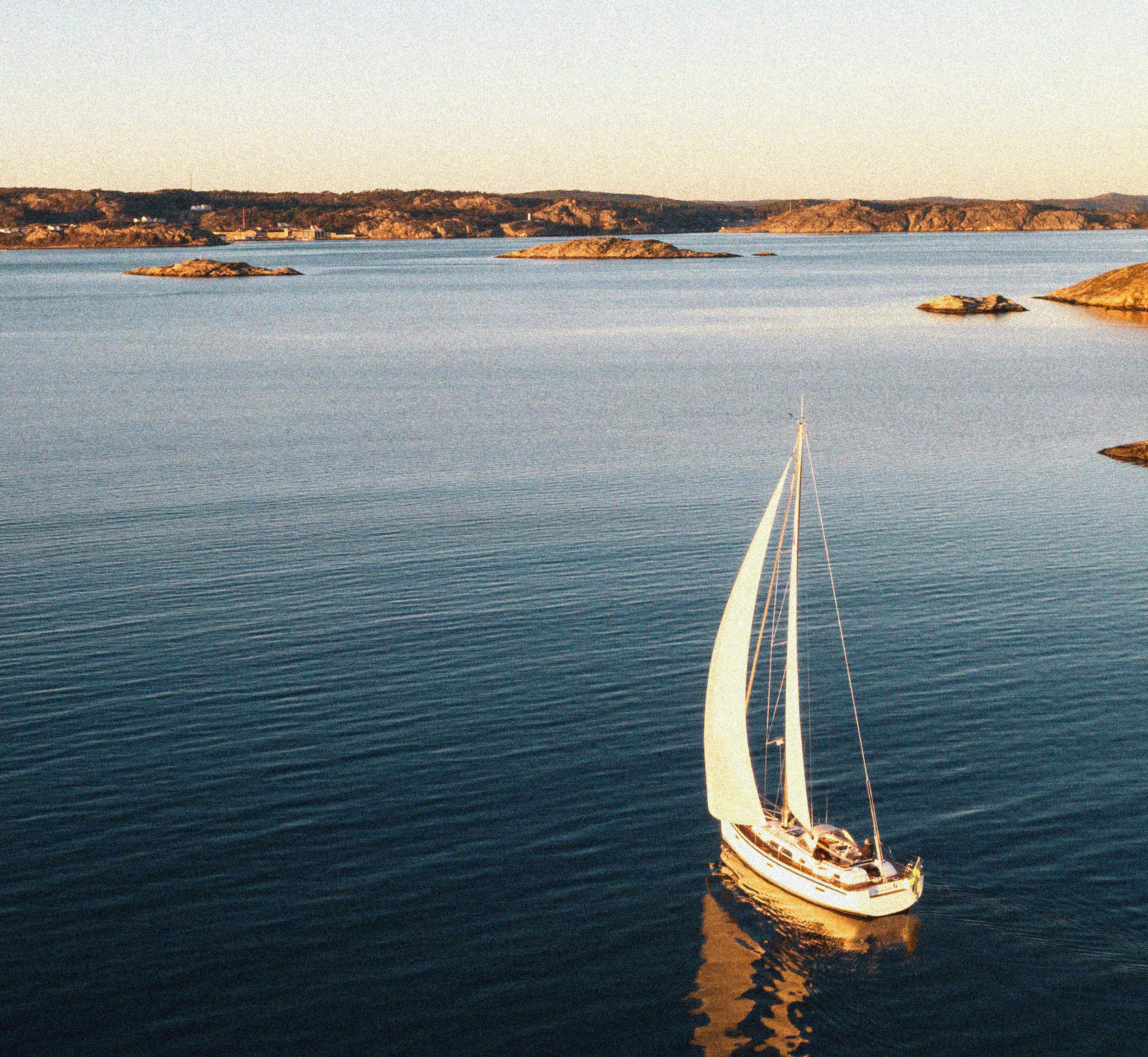 Velero blanco navegando en agua azul tranquila con islas rocosas en el fondo al atardecer.