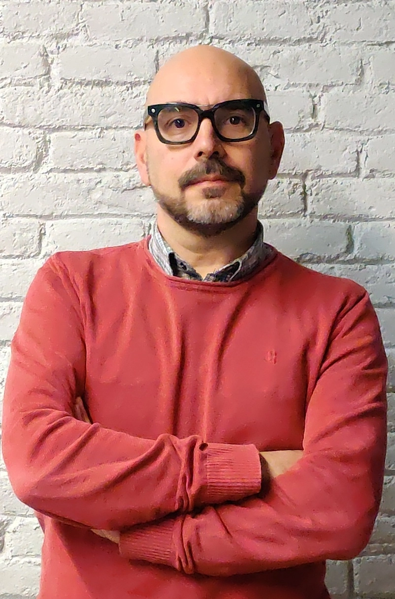 Hombre calvo con gafas negras y barba, vistiendo un suéter rojo y camisa a cuadros, posando frente a una pared de ladrillos blanca con los brazos cruzados.