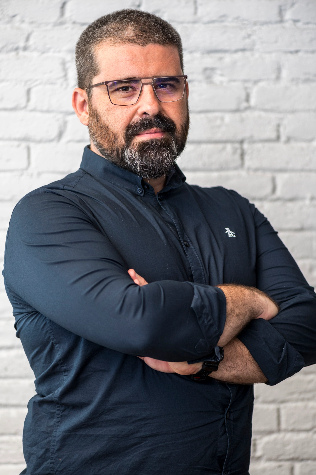 Hombre con gafas y barba entrecruzando los brazos frente a una pared de ladrillos blancos.
