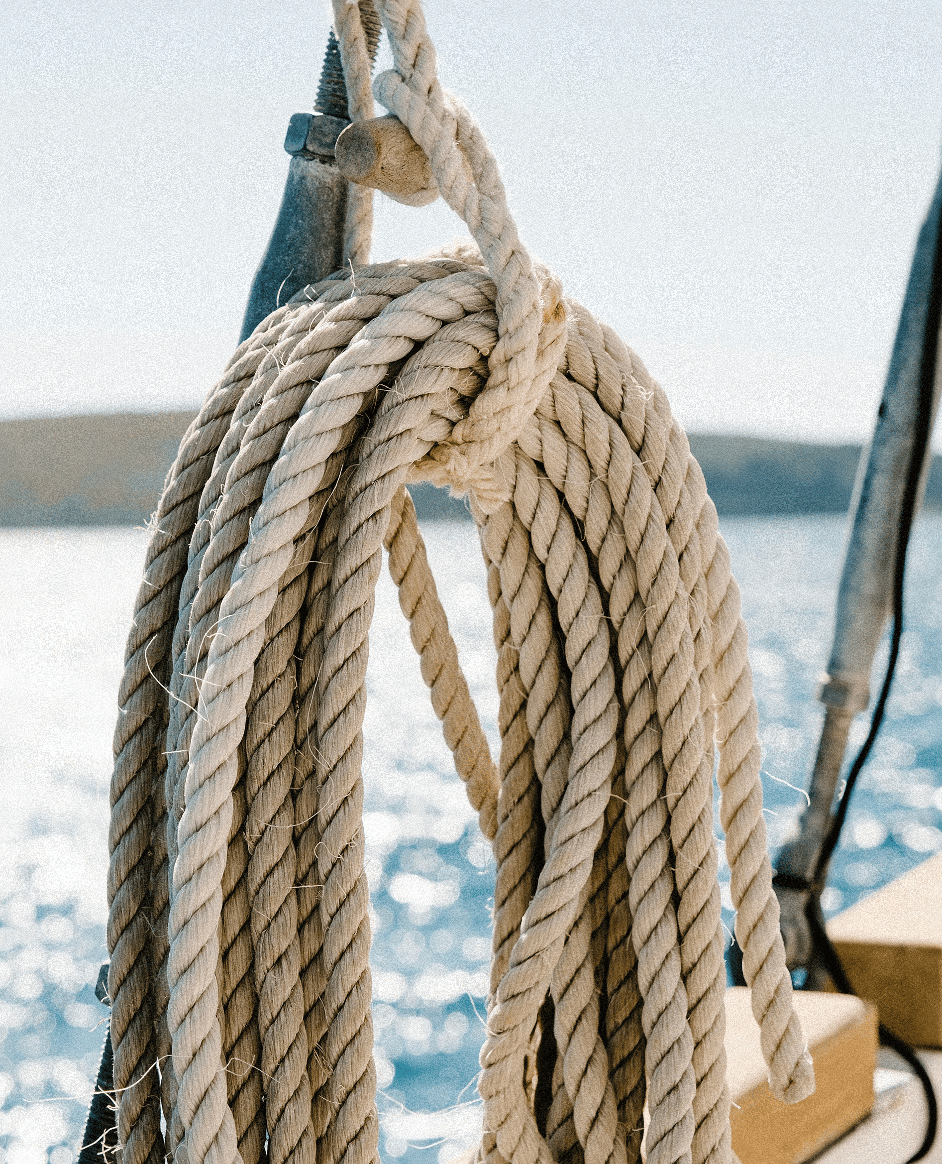 Cuerda gruesa enrollada suspendida en un barco con el mar brillante y un paisaje montañoso desenfocado al fondo.