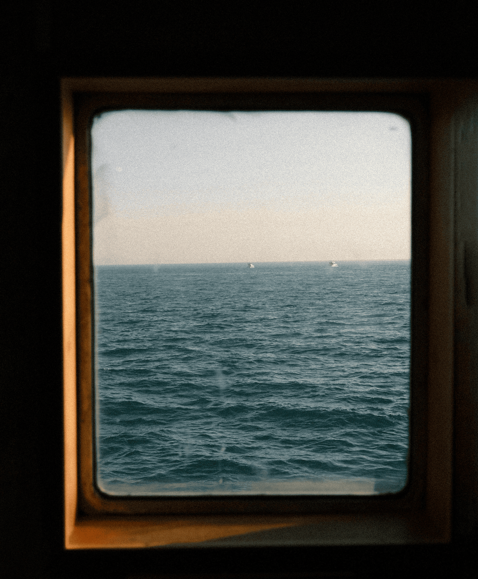Vista del mar con dos barcos pequeños en el horizonte, visto a través de una ventana con marco de madera.
