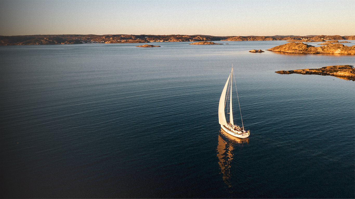 Velero blanco navegando en aguas tranquilas con islas rocosas al fondo durante el atardecer.