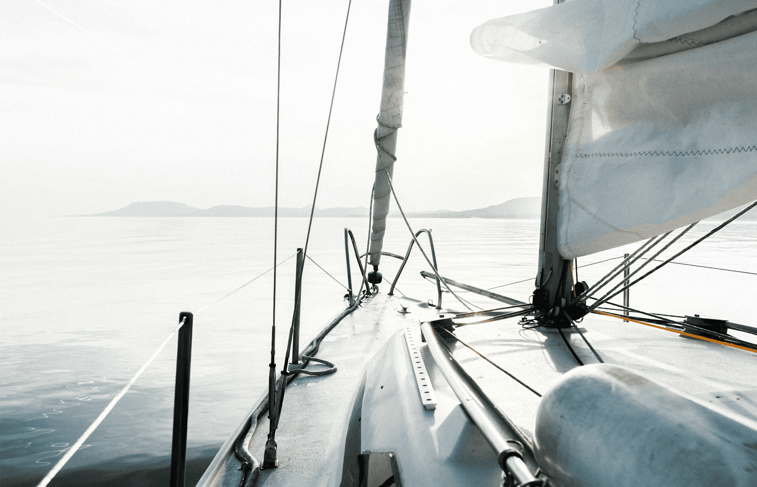 Vista desde el interior de un velero con la vela parcialmente desplegada navegando sobre mar tranquilo con montañas borrosas al fondo.