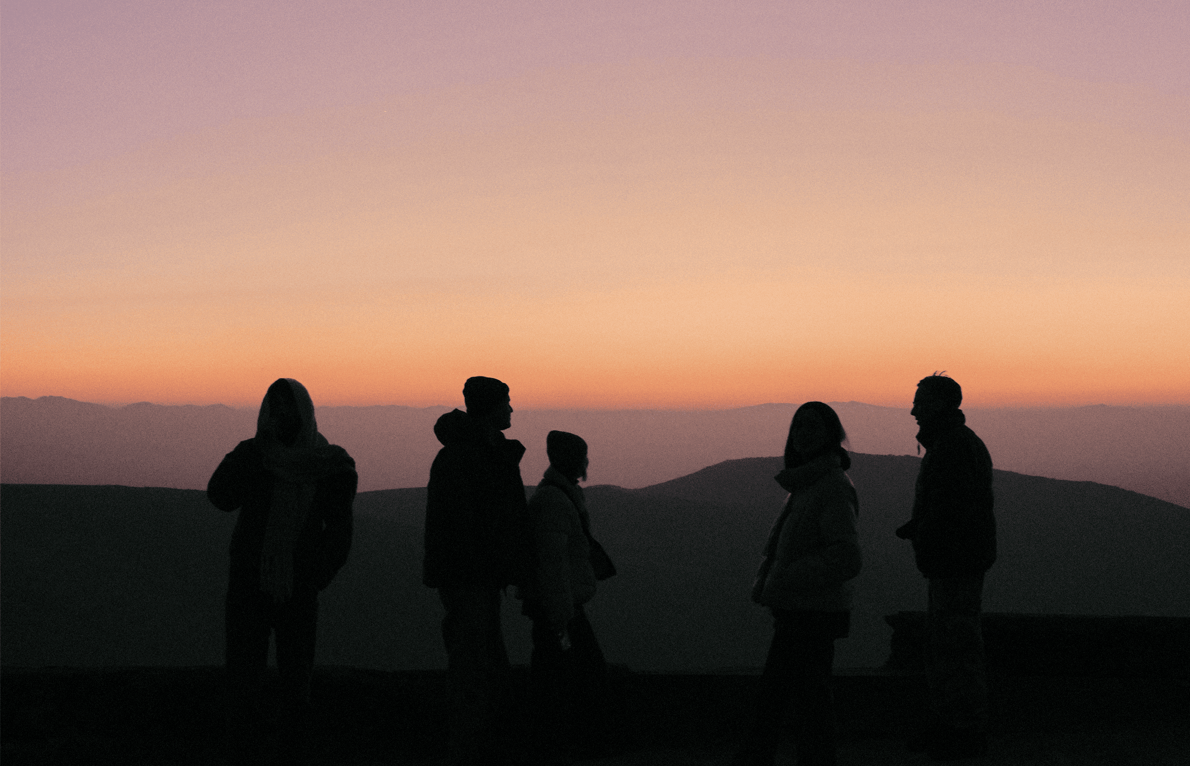 Siluetas de cinco personas de pie al aire libre con un atardecer de tonos anaranjados y púrpuras detrás de montañas.