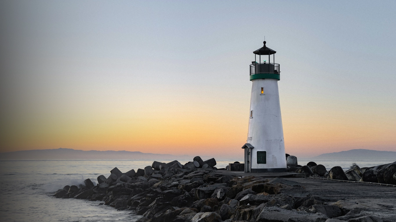 Faro blanco en un rompeolas de rocas durante el atardecer con el cielo en tonos naranja y azul.
