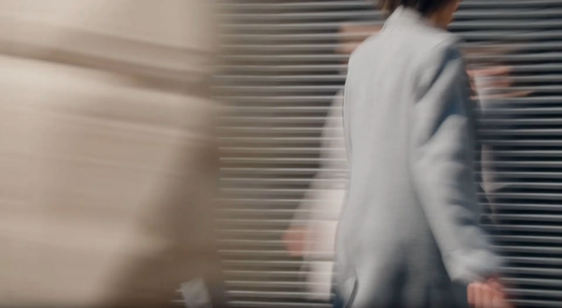 Blurred image of two people walking past each other in an urban setting with horizontal metal shutters in the background.