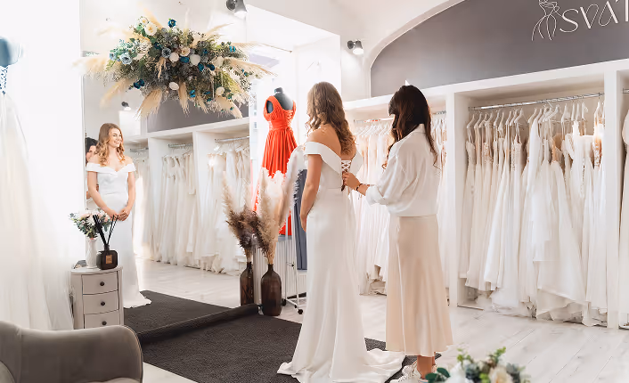 Žena si ve svatebním salonu zkouší bílý svatební šaty, zatímco ji druhá žena pomáhá zavázat šněrování na zádech.