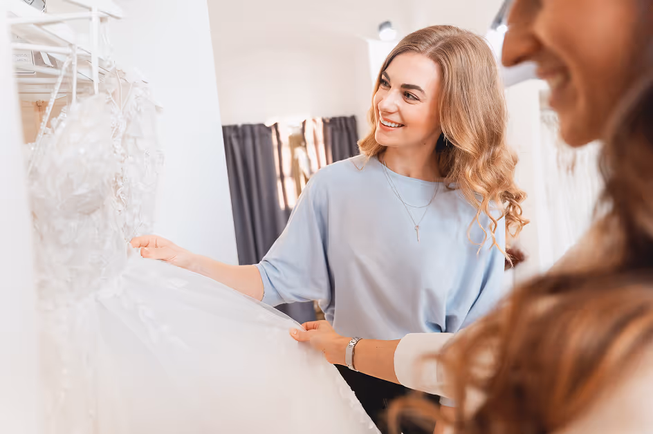 Dvě usměvavé ženy zkouší a prohlížejí si bílý svatební šaty v obchodě.
