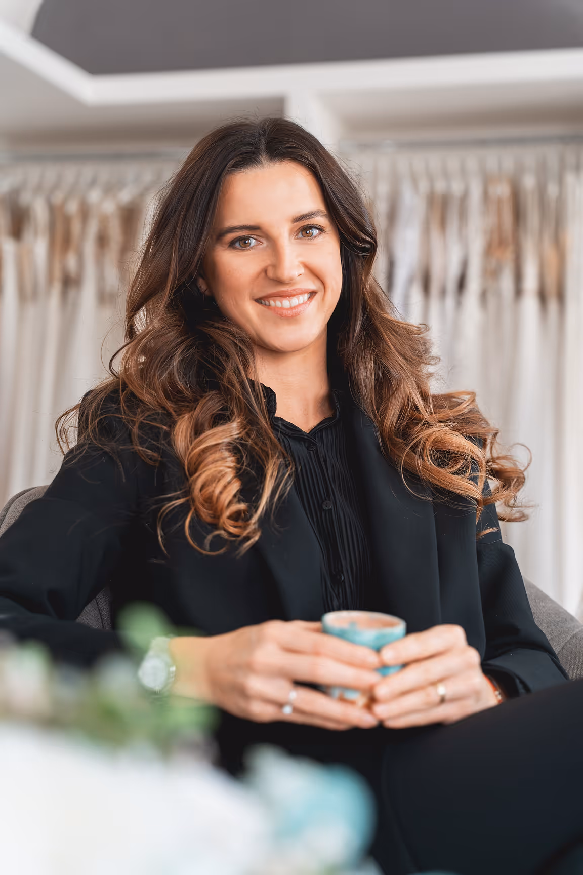Usměvavá žena s dlouhými vlnitými vlasy v černém oblečení sedí v křesle a drží modrý šálek.