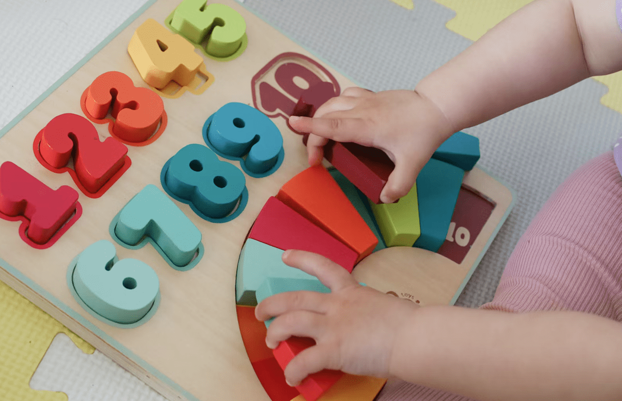 Child's hands arranging colorful wooden puzzle pieces including numbers and a rainbow on a play mat.
