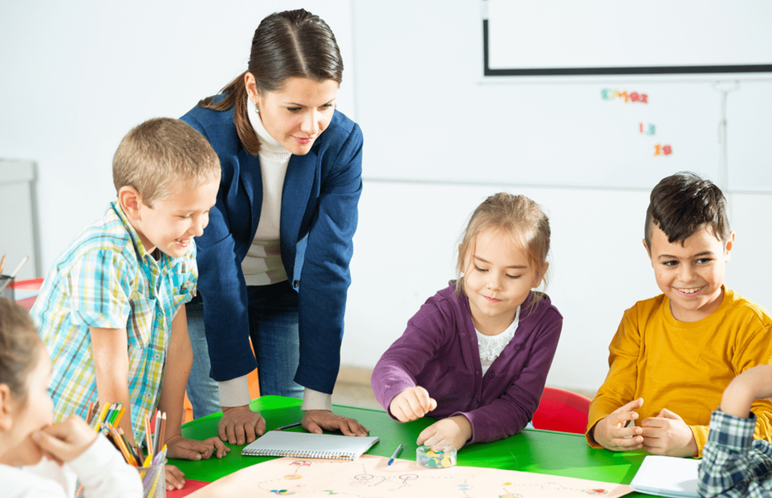Teacher and four children gathered around a green table, engaged in a group activity with colorful objects and drawings.