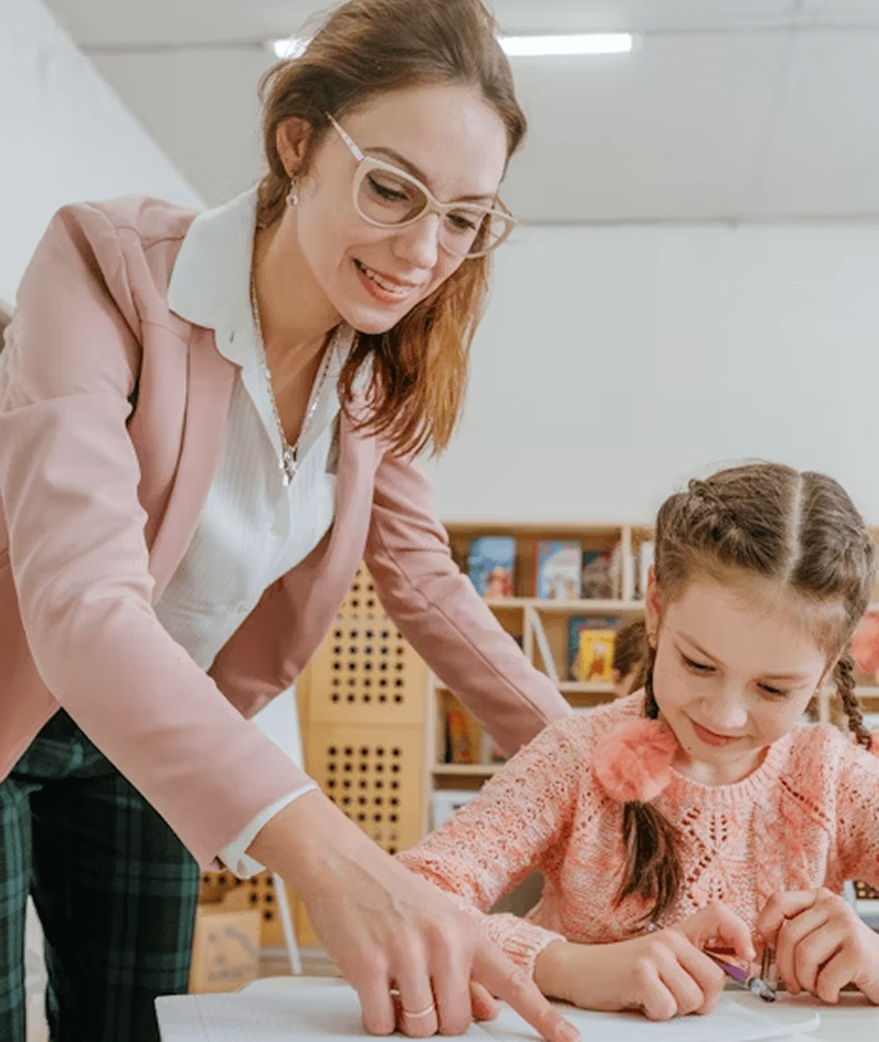 Teacher with glasses helping a young girl with braids working on a project at a table in a classroom.