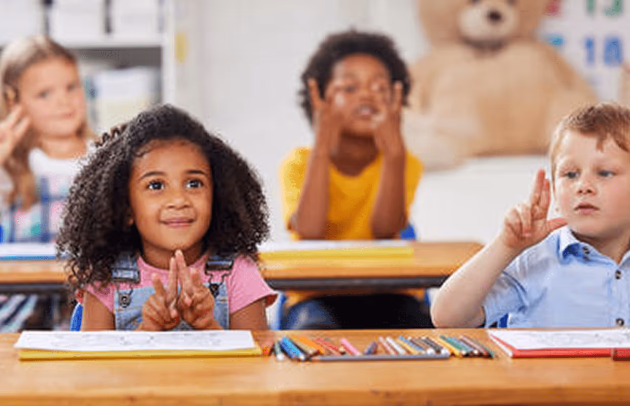 Group of young children sitting at desks in a classroom, holding up fingers to count or signal numbers.