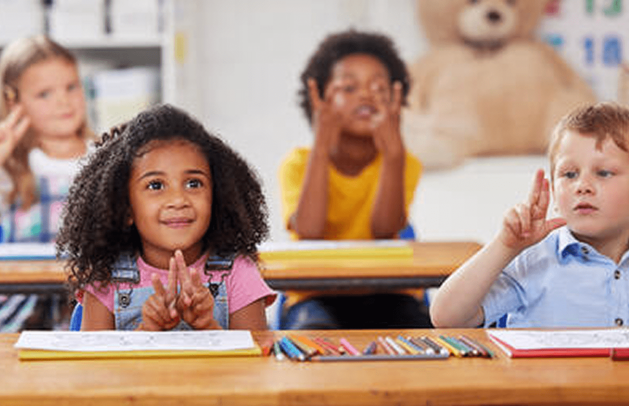 Group of young children sitting at desks in a classroom, holding up fingers to count or signal numbers.
