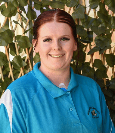 Miss Kesiah wearing a blue polo shirt standing in front of leafy green plants.