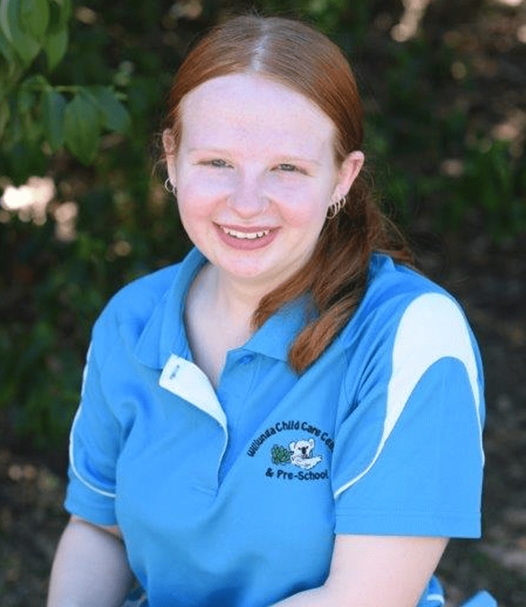 Smiling Miss Katlyn with red hair wearing a blue polo shirt with a child care center logo, sitting outdoors in front of greenery.
