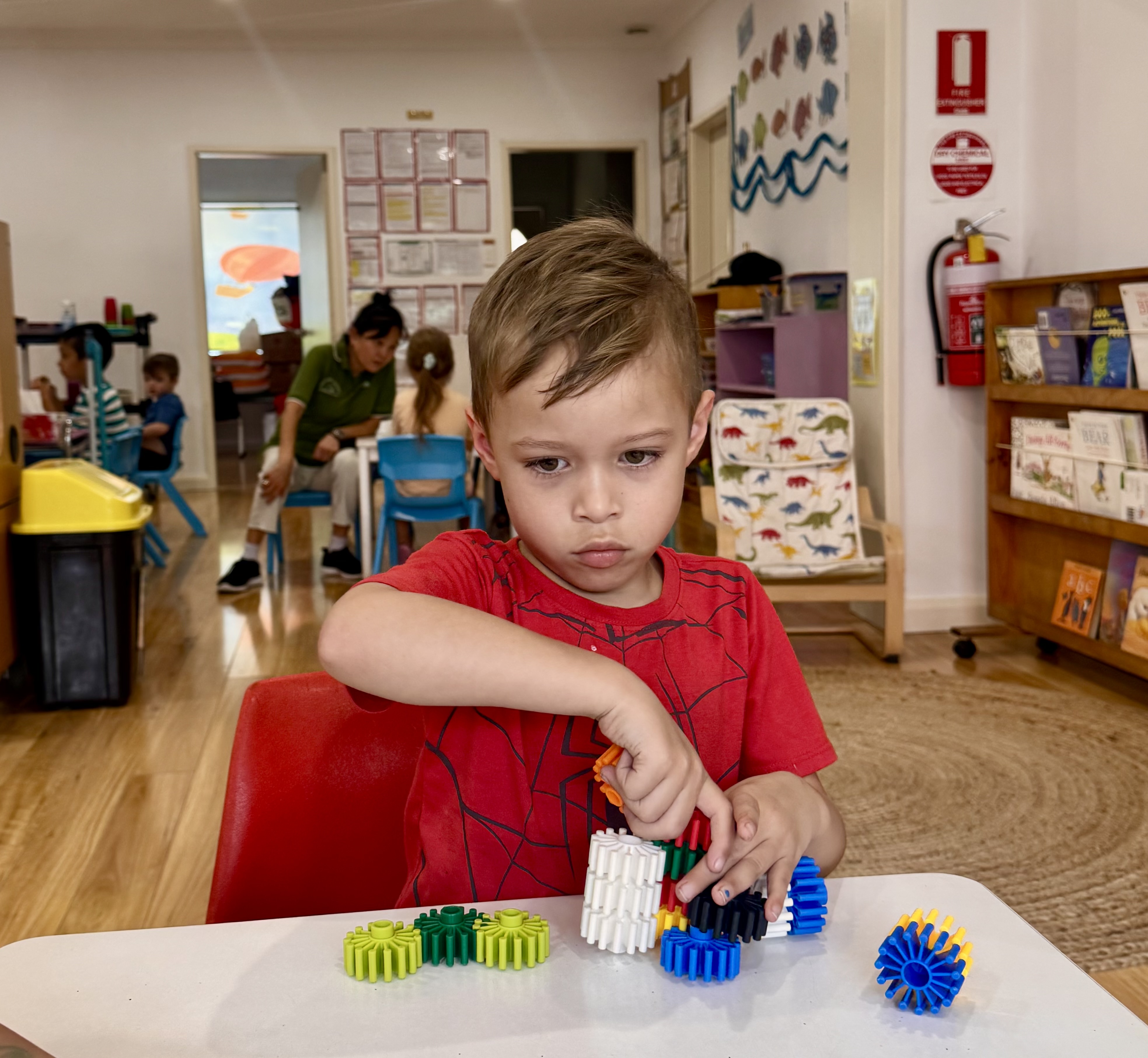 Young boy in a red spider web shirt focused on assembling colorful plastic gears at a table in a classroom. A student from Quakers Hill Kindergarten & Child Care Centre.