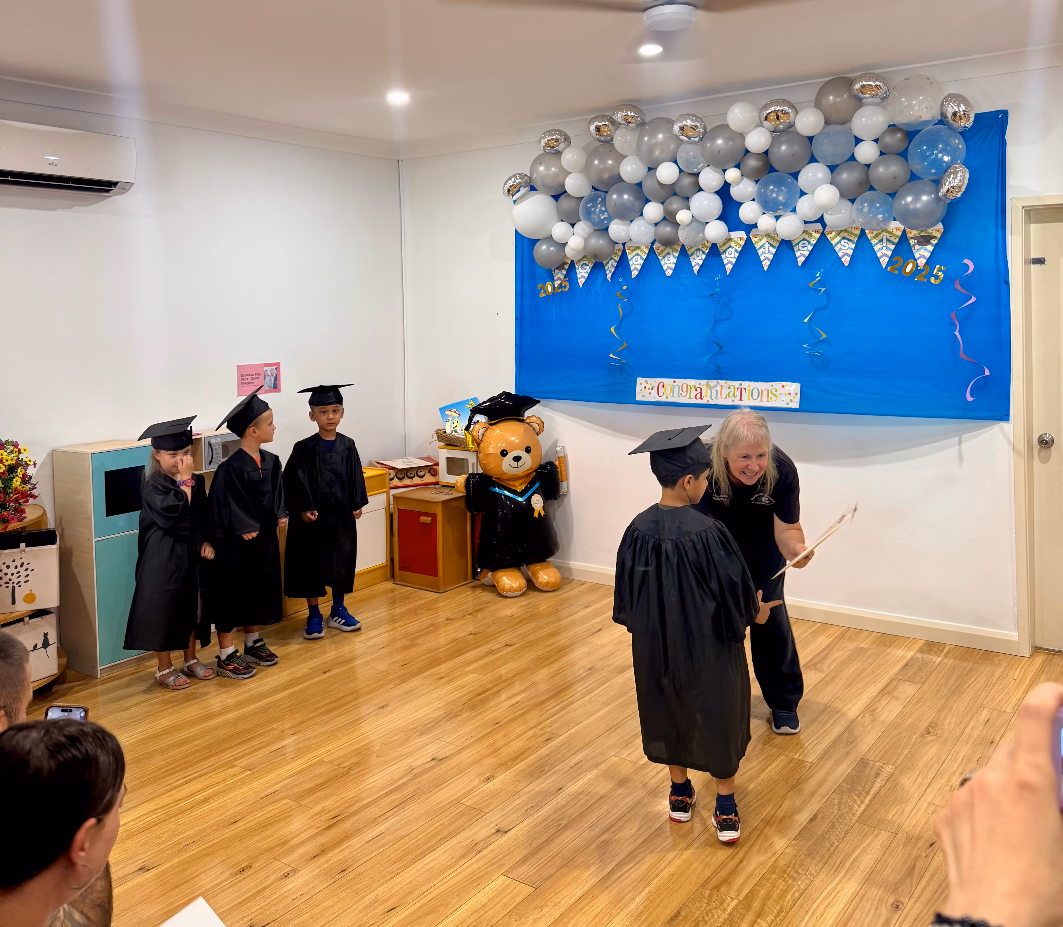 Young children in graduation caps and gowns at a kindergarten graduation ceremony receiving certificates, with balloon decorations and a congratulatory banner.