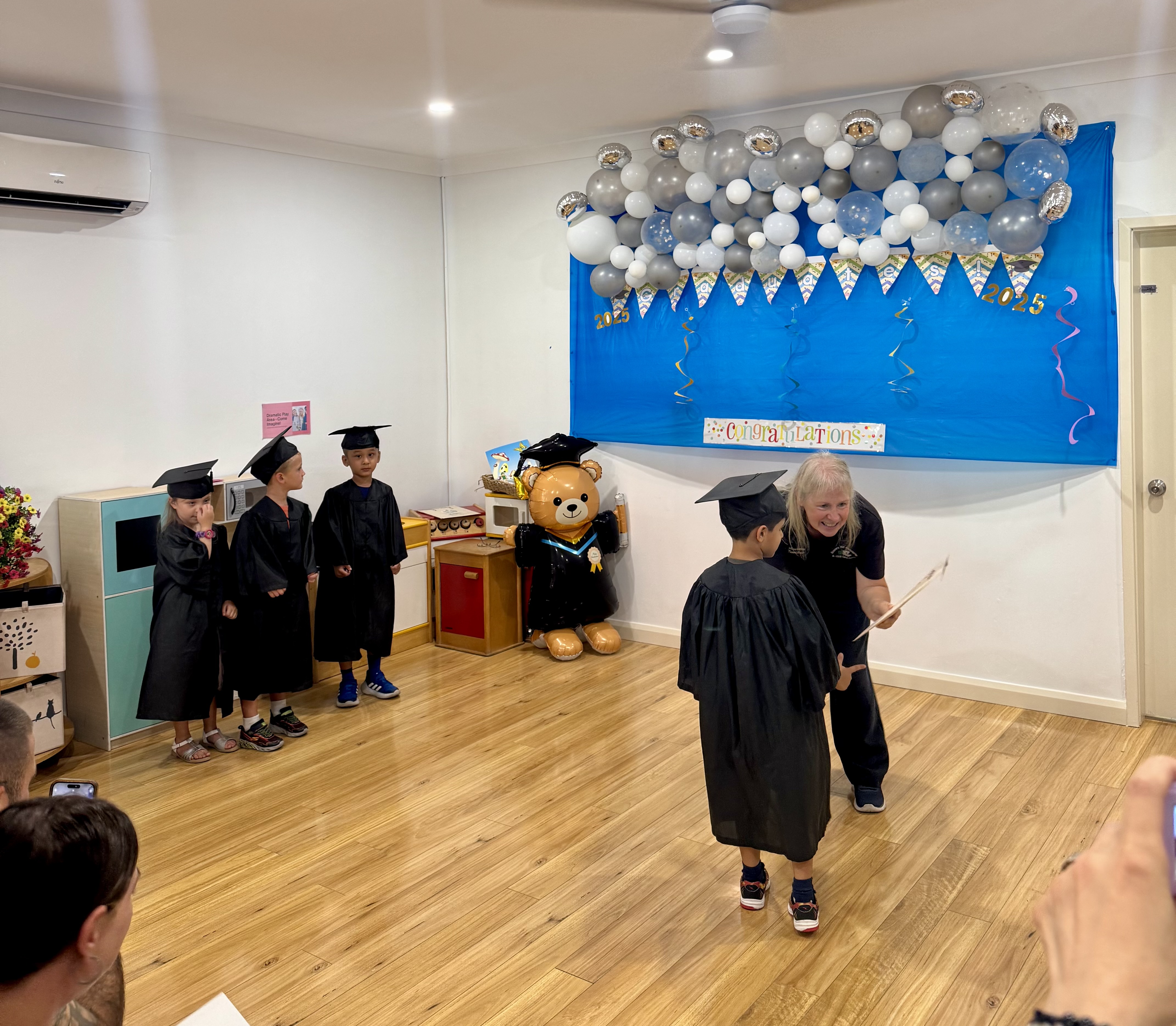 Young children in graduation caps and gowns at a kindergarten graduation ceremony receiving certificates, with balloon decorations and a congratulatory banner.