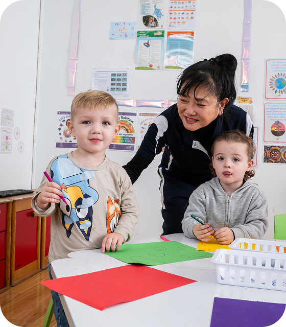 Young girl coloring a picture with colored pencils at a classroom table with a boy and educational posters in the background.