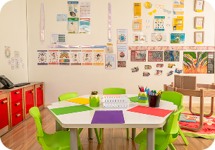 Young girl coloring a picture with colored pencils at a classroom table with a boy and educational posters in the background.
