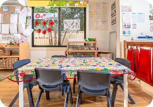 Young girl coloring a picture with colored pencils at a classroom table with a boy and educational posters in the background.