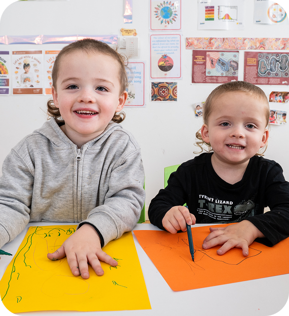 Young girl coloring a picture with colored pencils at a classroom table with a boy and educational posters in the background.