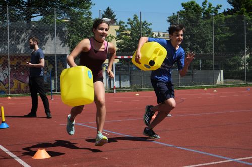 Two teenagers running on a sports track carrying large yellow containers in order to raise money for water.