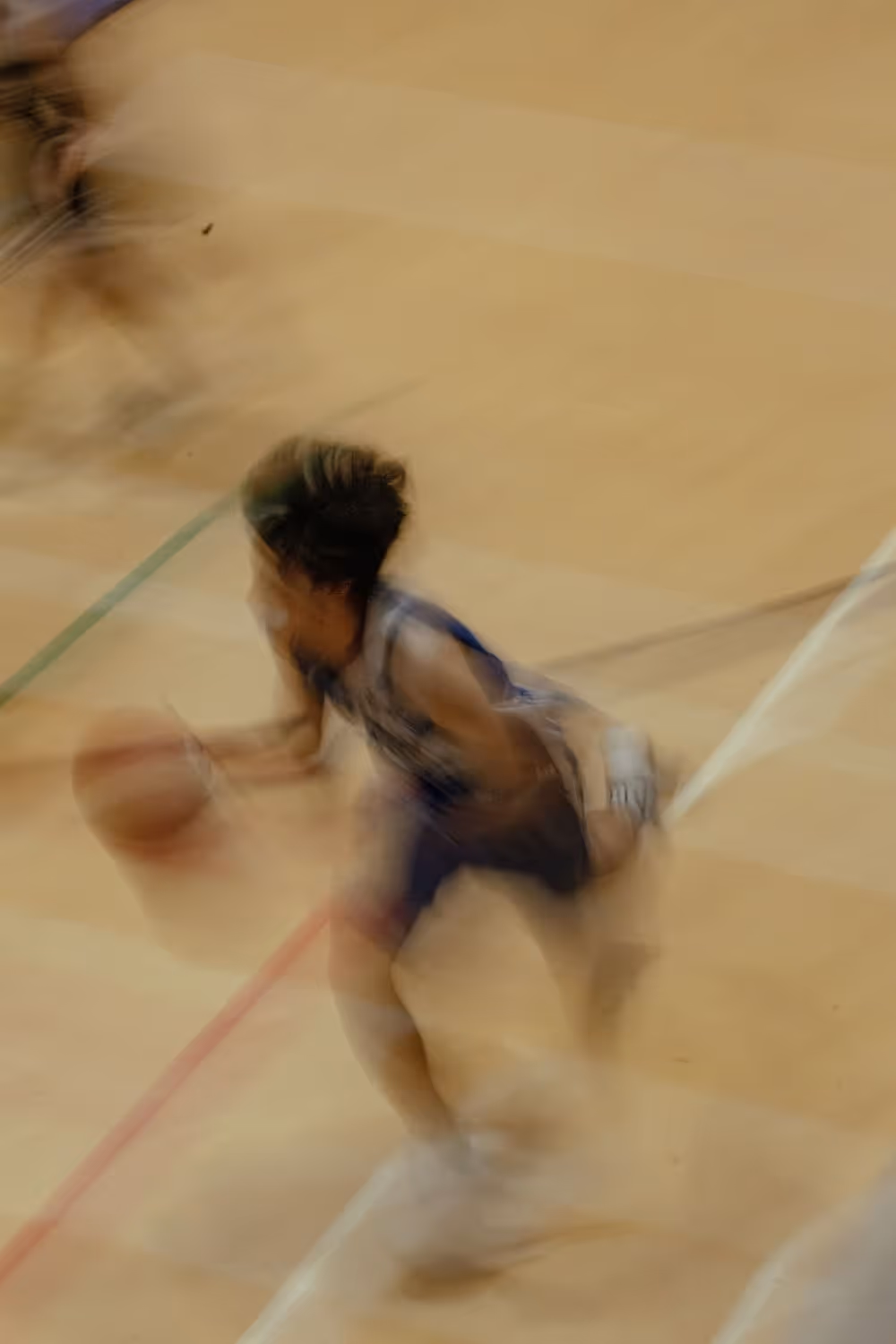 Joueur de basket en mouvement flou sur un terrain intérieur, dribblant rapidement le ballon. L’effet de flou accentue la vitesse et l’intensité de l’action.