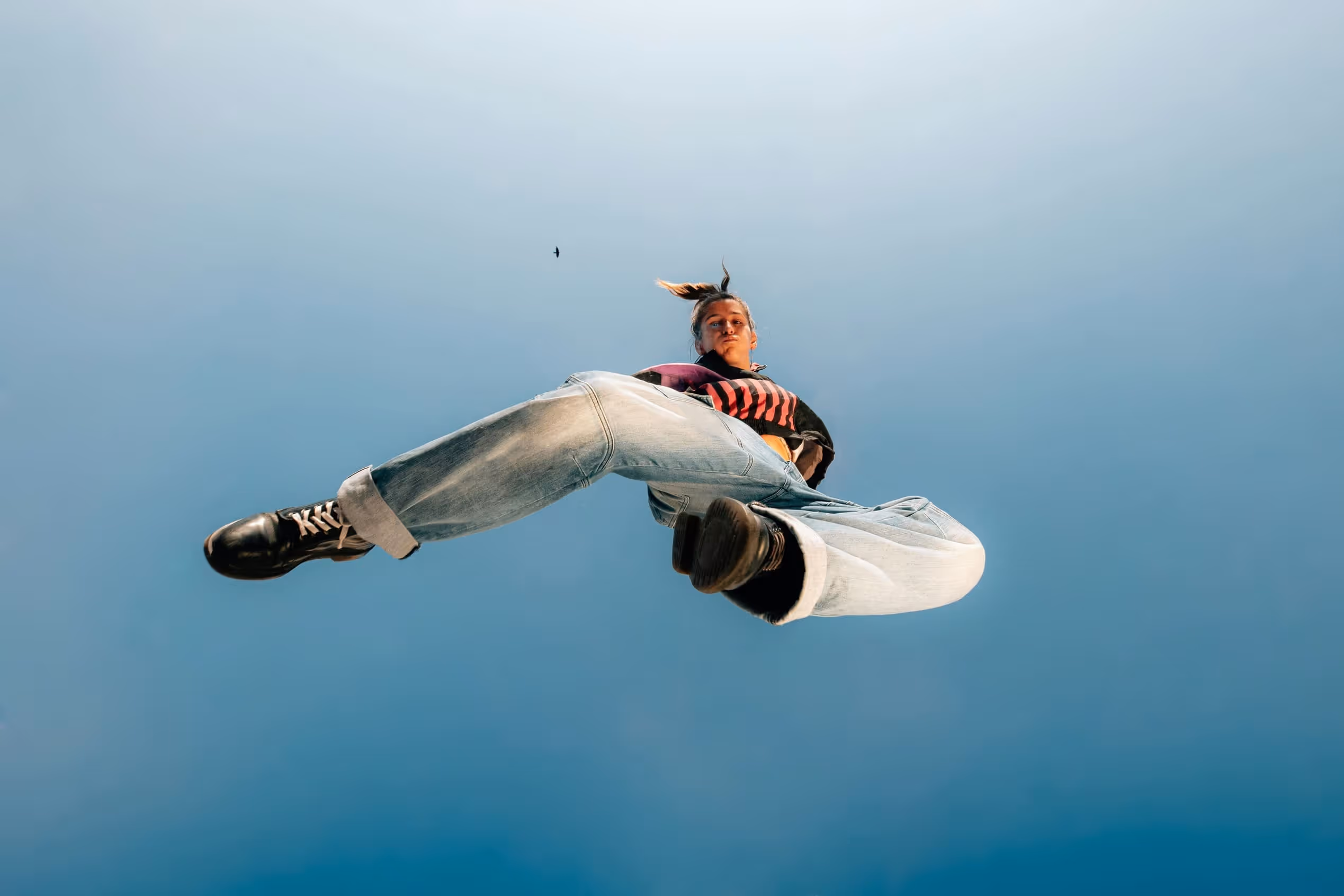 Une jeune femme photographiée en contre-plongée en plein saut, habillée en jean large, t-shirt noir et baskets noires et roses, avec le ciel bleu en arrière-plan. L’image capture une sensation de liberté et de mouvement.