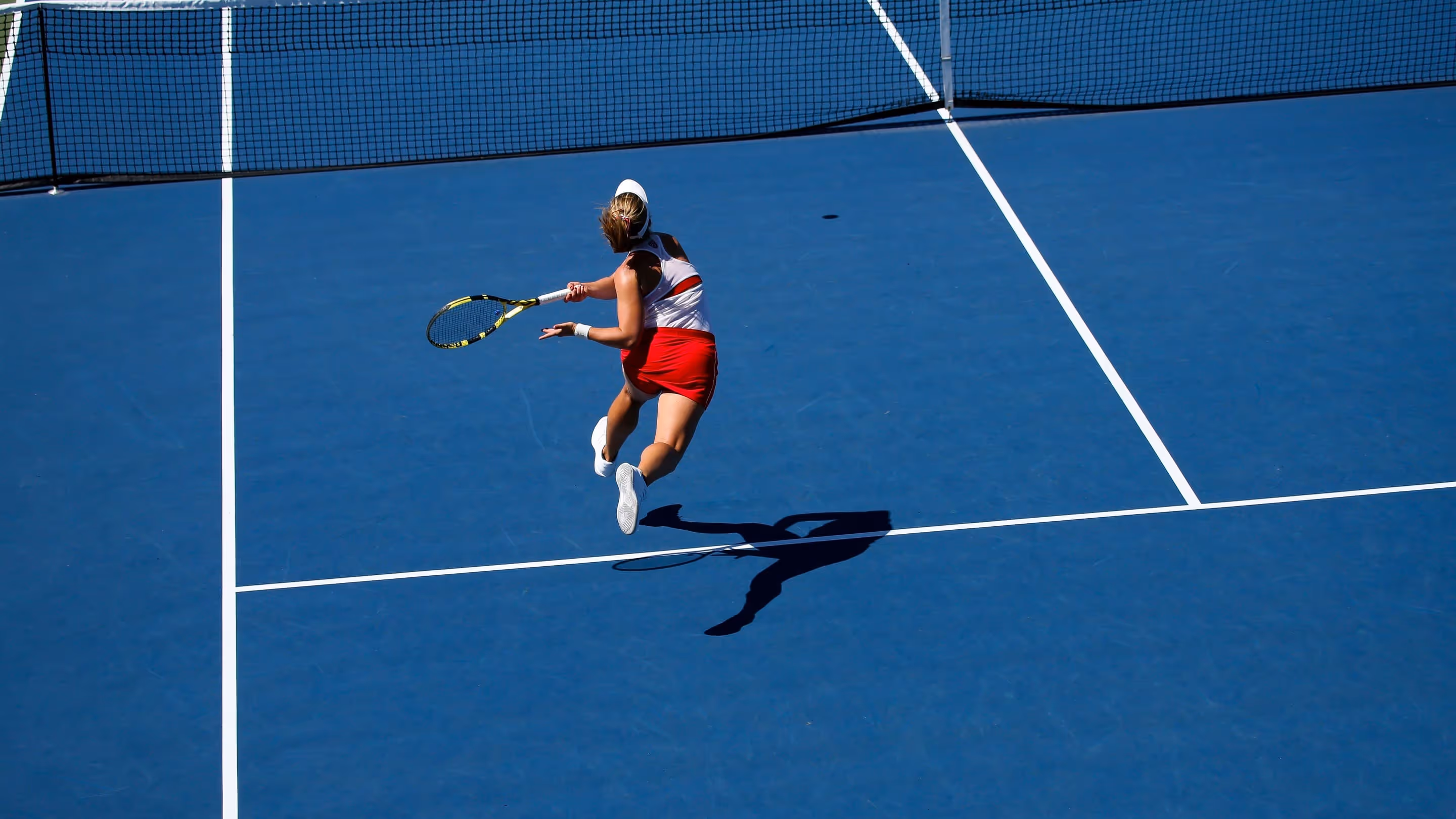Joueuse de tennis en action sur un court bleu, effectuant un coup de raquette avec intensité pendant un match. Son ombre projetée au sol souligne le dynamisme de la scène.