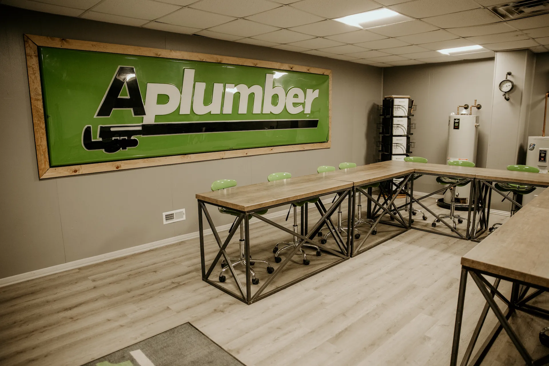 Modern conference room with wooden tables arranged in a U-shape, green chairs on wheels, and a green sign on the wall that reads 'A plumber'.
