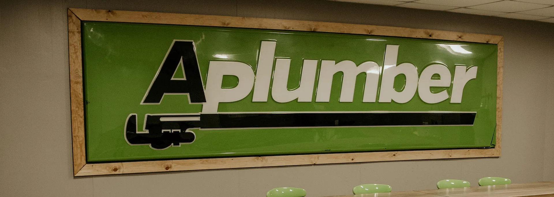 Green sign with wooden frame displaying the logo text 'Aplumber' with a black pipe wrench graphic below.