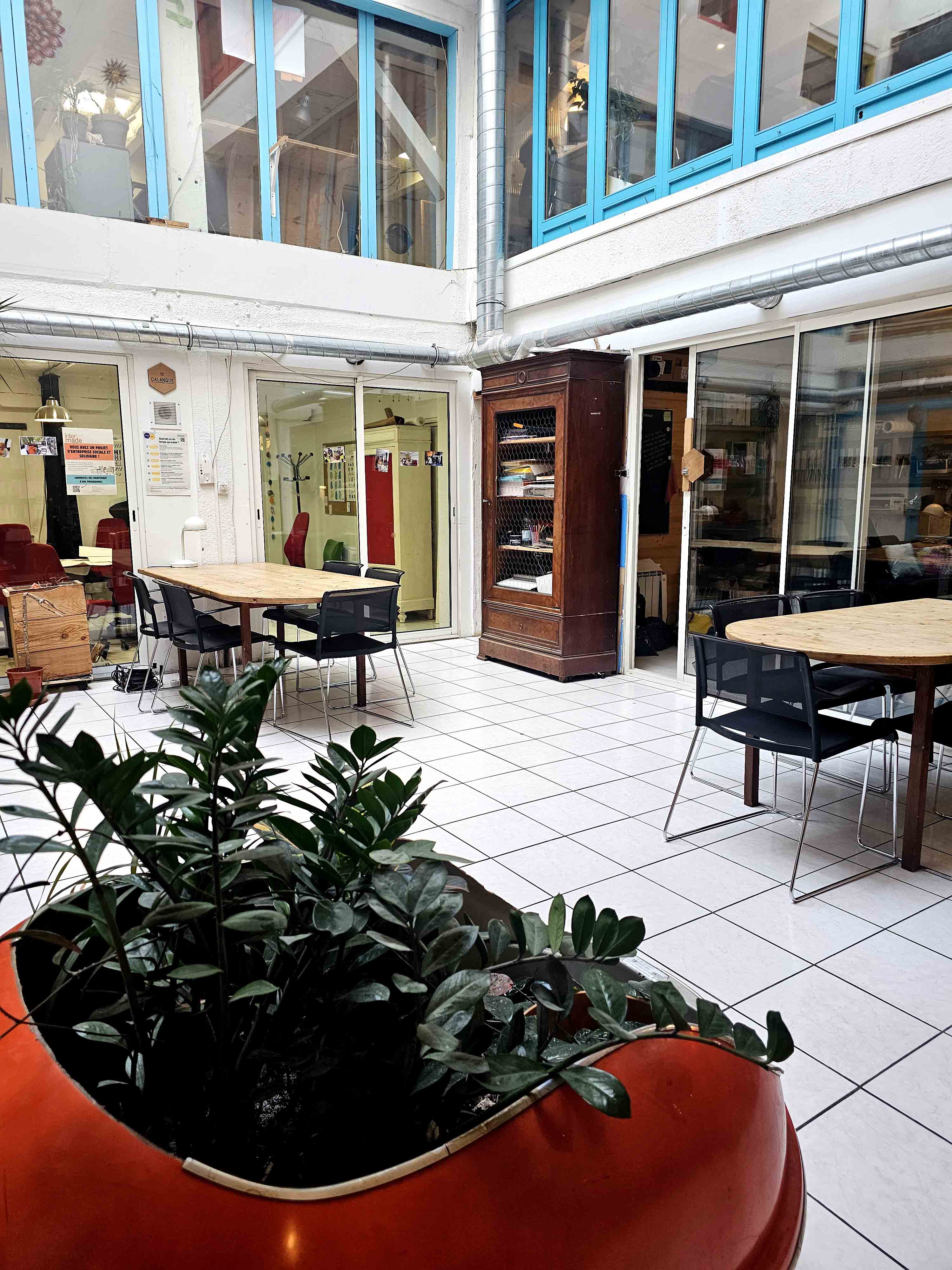 Espace commun lumineux avec grandes fenêtres bleu ciel, deux tables en bois entourées de chaises noires, plante verte dans un grand pot rouge et sol carrelé blanc.
