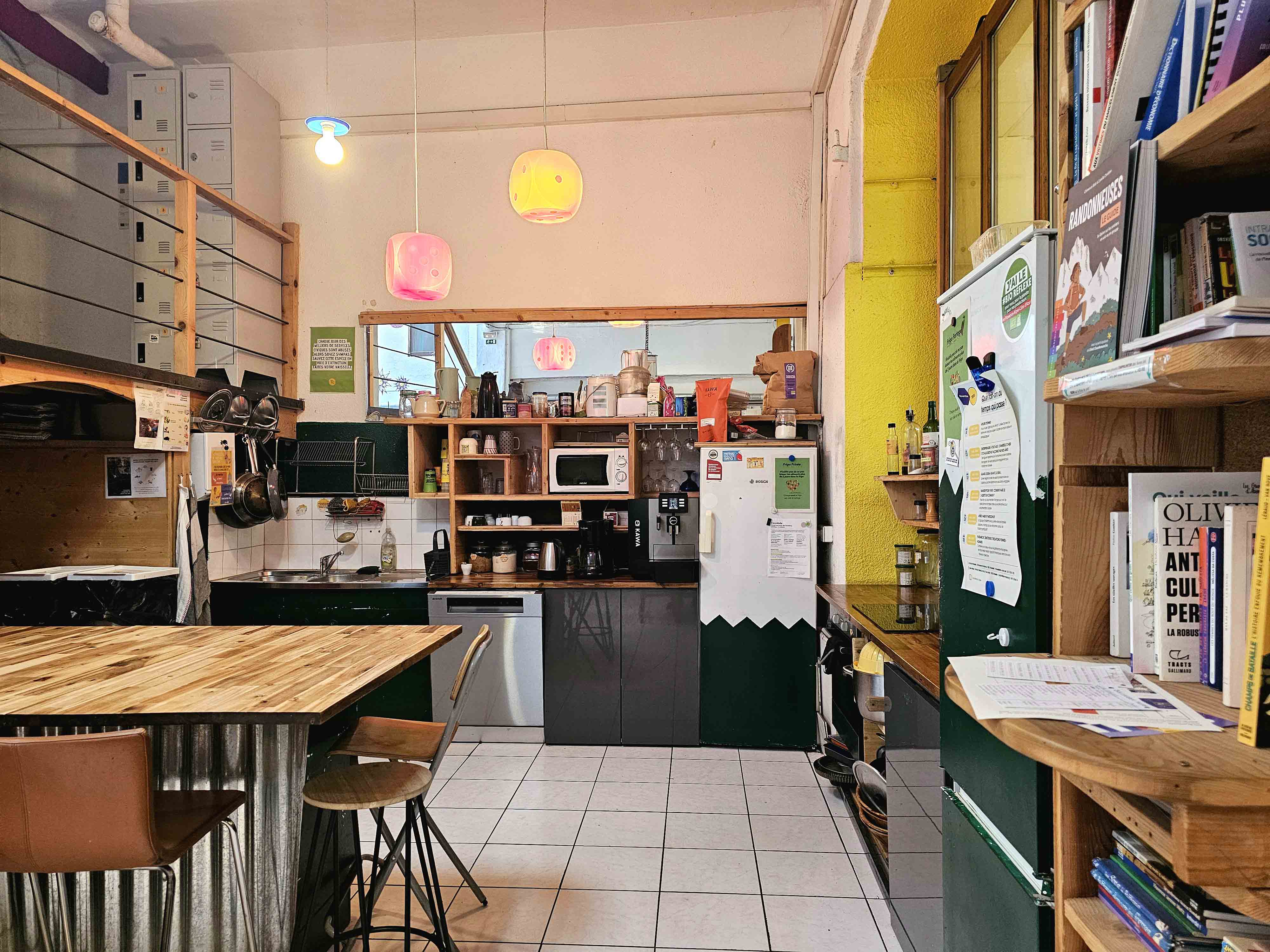 Cuisine communautaire avec comptoir en bois, tabourets, réfrigérateur blanc, micro-ondes, cafetière, étagères remplies d'objets et livres sur le côté droit.