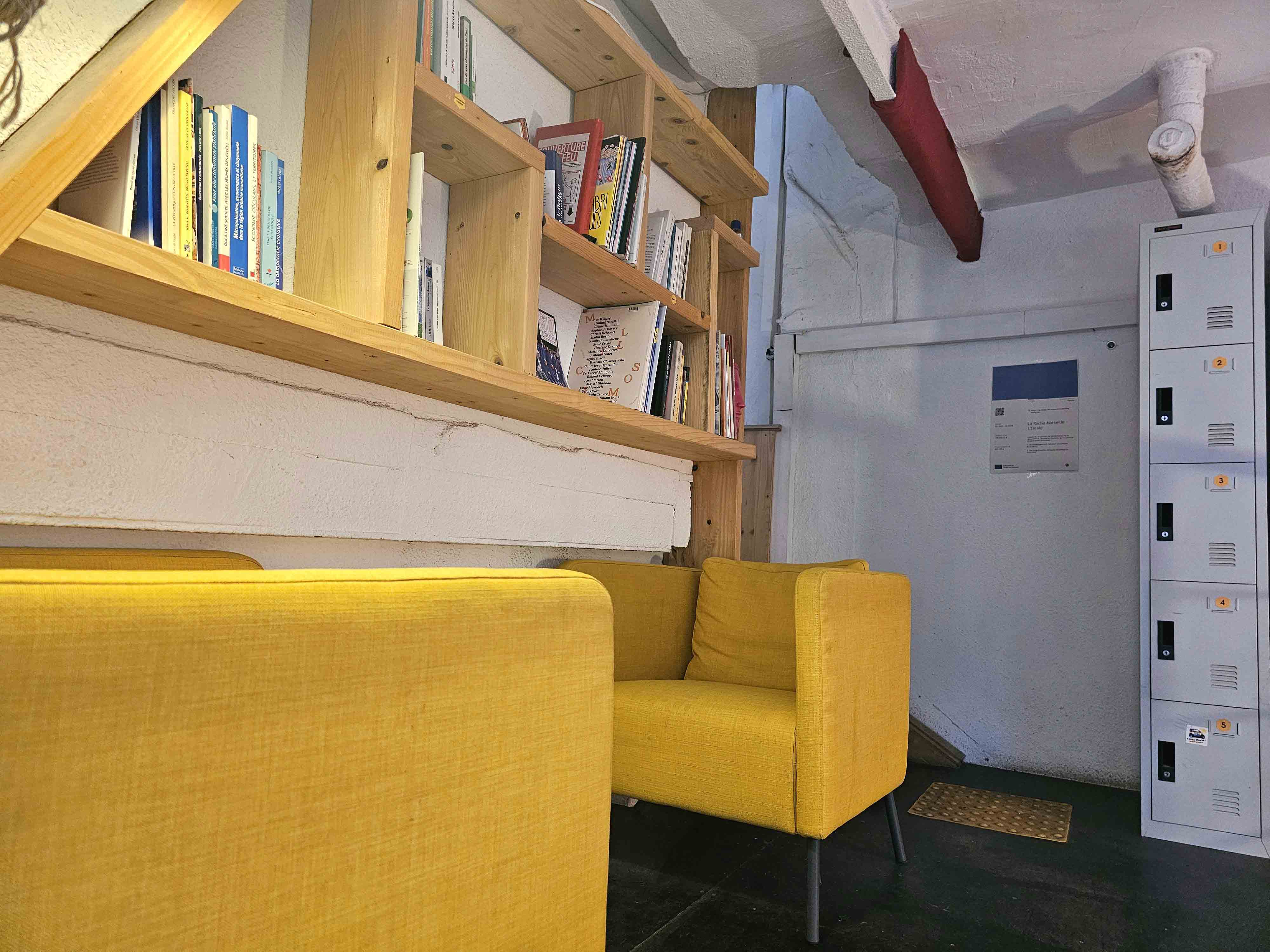 Coin lecture avec canapés jaunes, étagère en bois remplie de livres et casiers métalliques blancs sur un mur blanc.