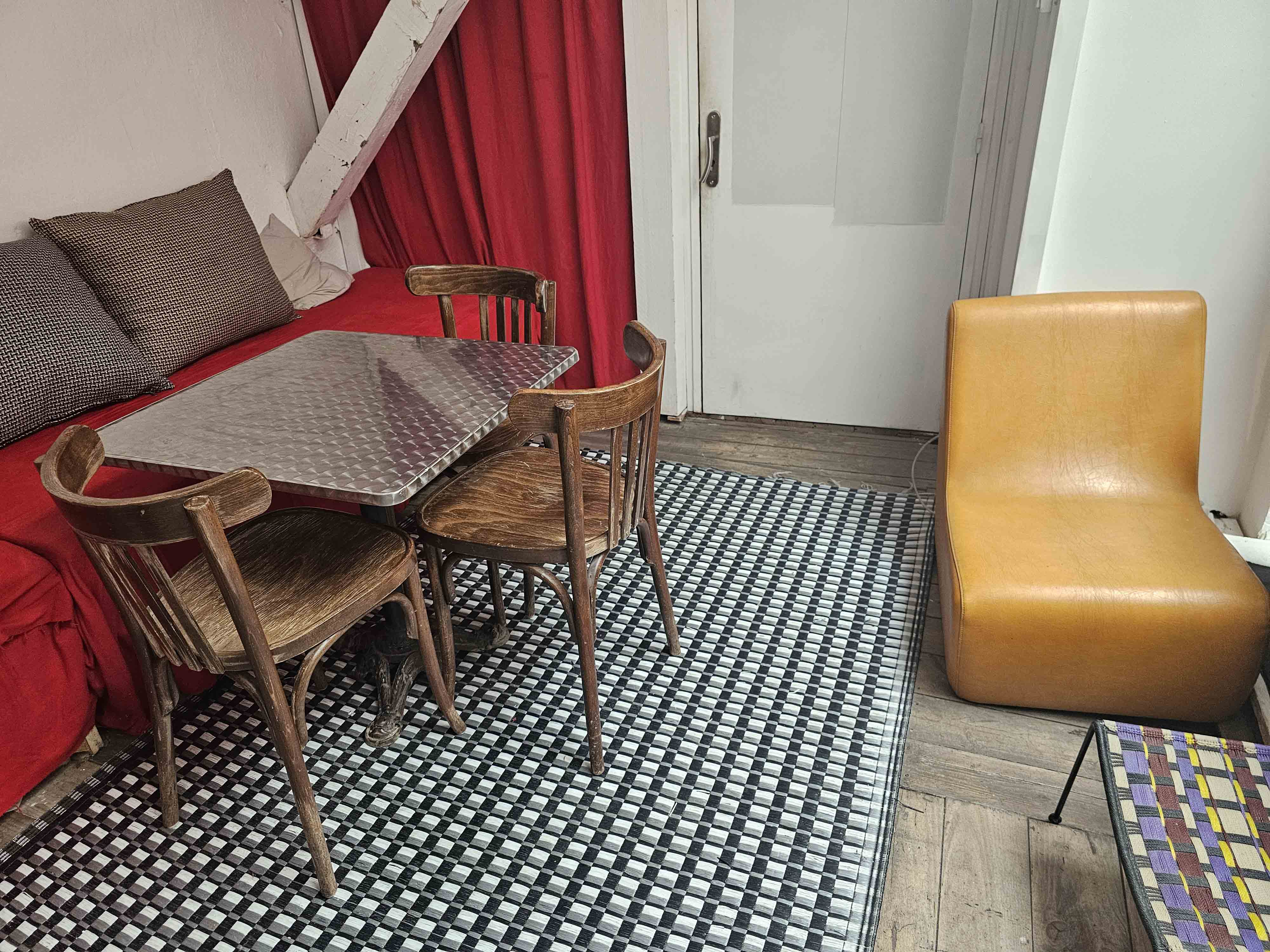 Coin terrasse avec table métallique, trois chaises en bois, un canapé rouge avec coussins et un fauteuil marron clair sur un tapis à carreaux noir et blanc.