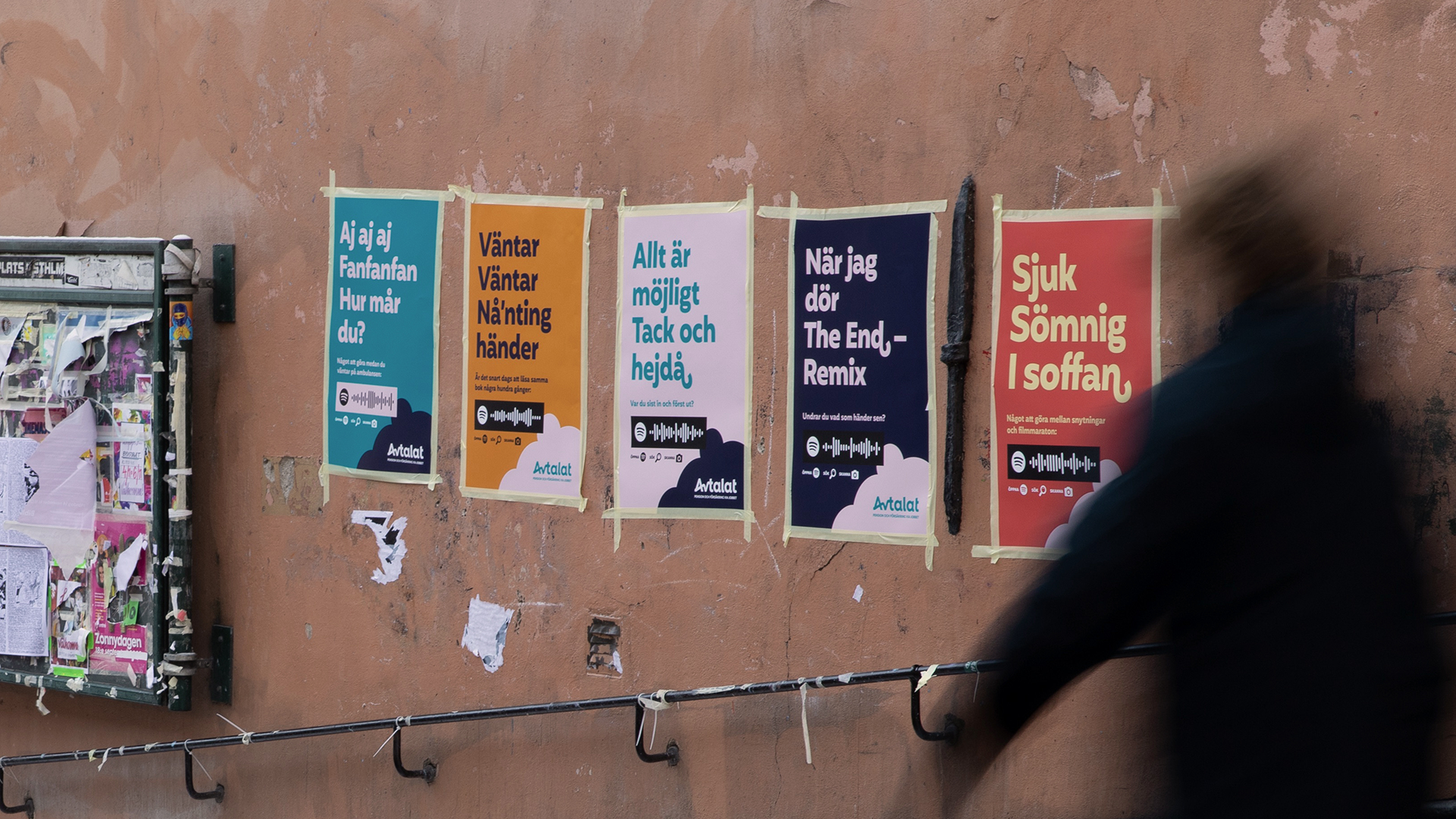 Fem färgglada affischer från Avtalat upptejpade på en putsad husvägg. Affischerna har olika rubriker som anspelar på låttexter och innehåller Spotify-koder. En suddig gestalt passerar i förgrunden, vilket ger en känsla av rörelse i stadsmiljön.
