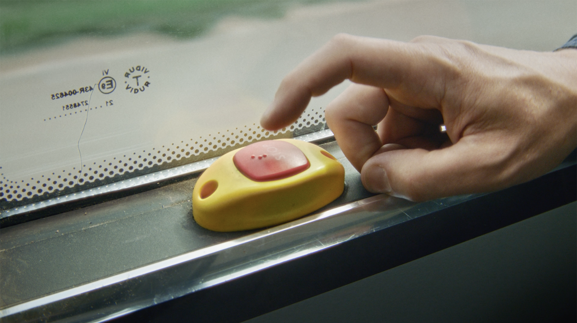 Närbild på en hand inne i en buss där ett finger precis ska trycka på en stoppknapp. Knappen är gul med en röd mittpunkt och är monterad på den mörka listen under ett fönster. 