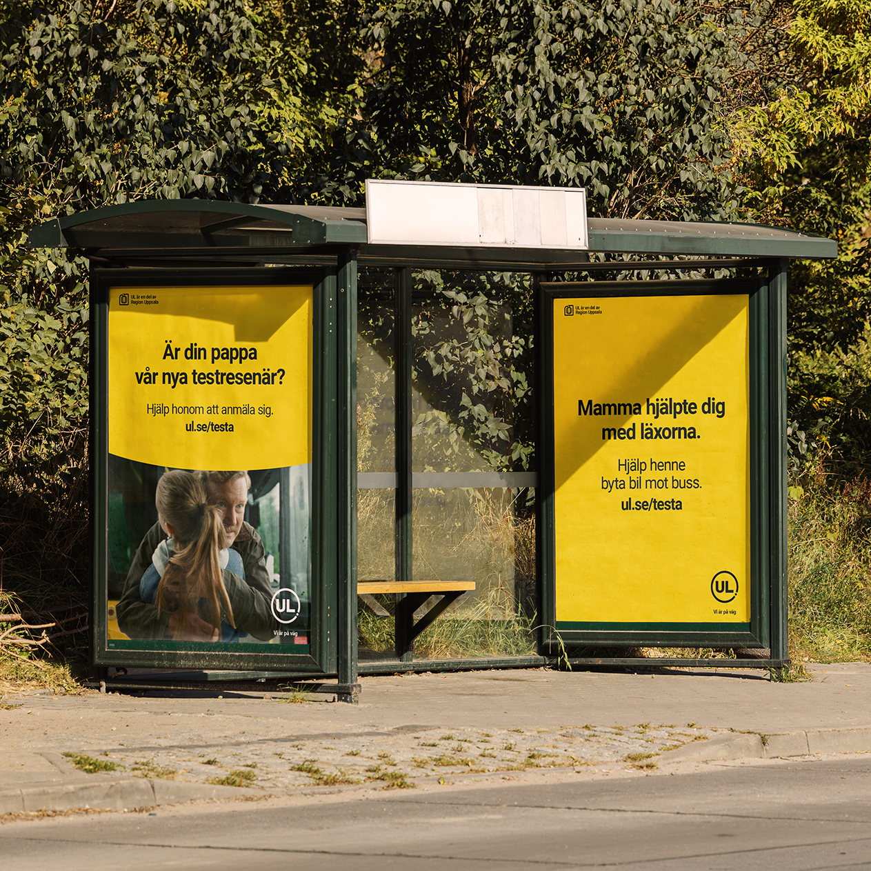 En busshållplats i profil med två gula reklamaffischer från UL (Kollektivtrafiken i Uppsala län). Hållplatsen är placerad utomhus med grönska i bakgrunden.