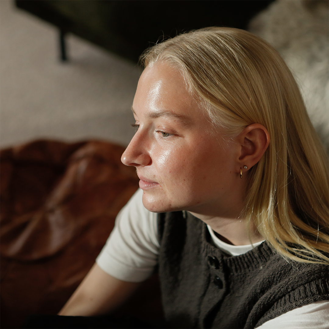 Porträtt av en ung kvinna med långt, ljusblont hår i trekvarts profil. Hon bär en vit t-shirt under en mörkgrå stickad väst och sitter inomhus med en suddig bakgrund.