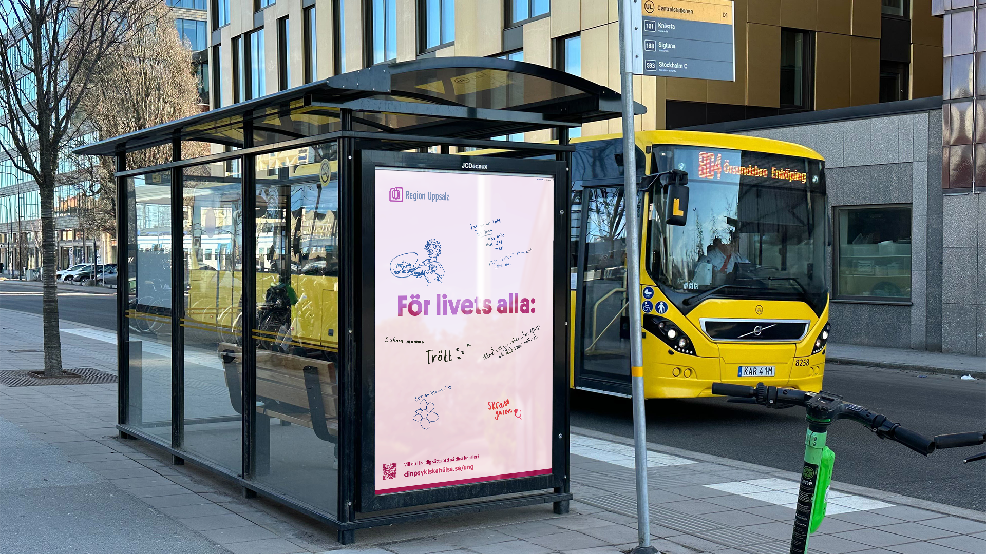 En ljusrosa reklampelare från Region Uppsala i ett modernt stadsområde, monterad på kortsidan av en busshållplats i glas. Affischen har rosa och svart text på svenska med budskapet "För livets alla:" omgiven av teckningar och handskrivna anteckningar.