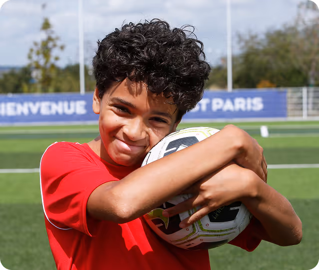 Smiling boy in red shirt hugging a soccer ball on a green soccer field.