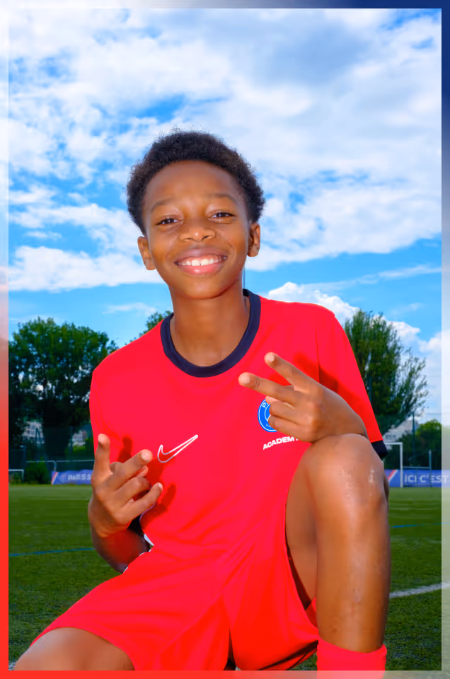 Smiling boy in red soccer uniform kneeling on a soccer field under a blue sky with clouds.