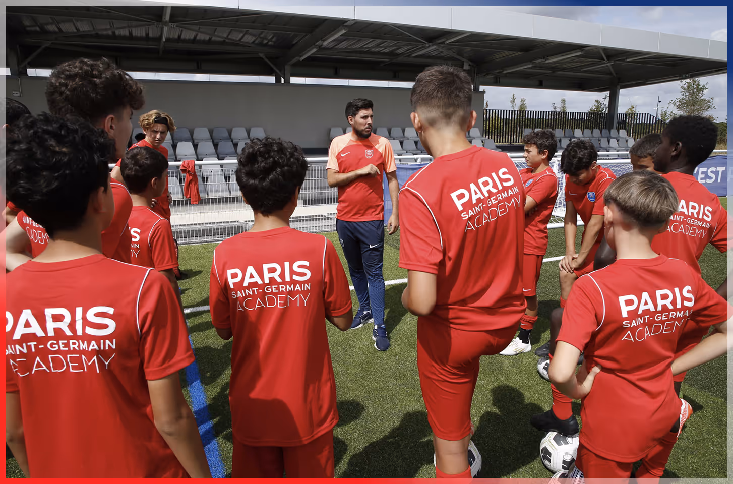 Coach giving instructions to young soccer players wearing red Paris Saint-Germain Academy uniforms on a soccer field.