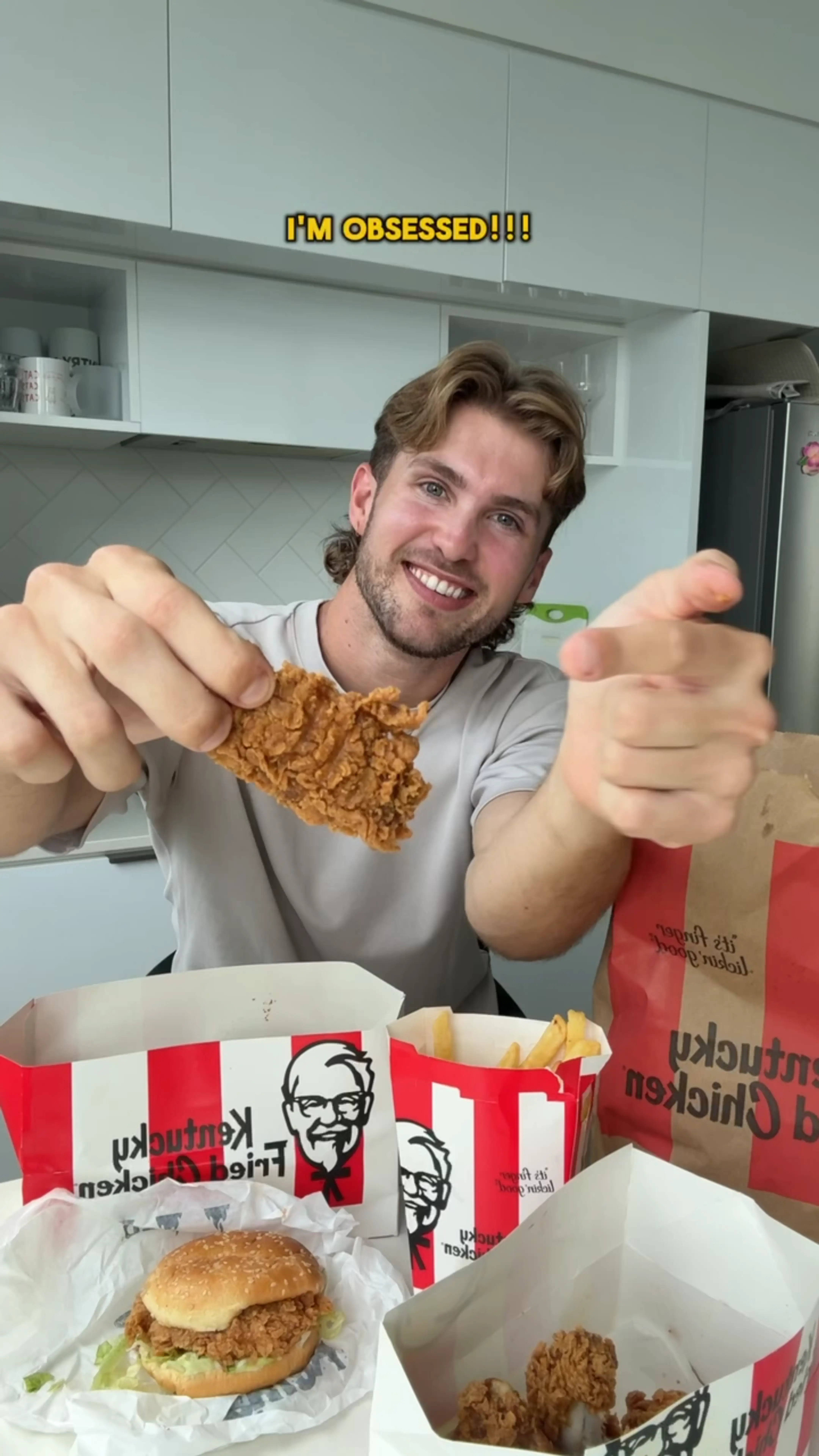 Smiling man holding a piece of fried chicken with boxes of KFC chicken, fries, and a chicken sandwich on the table in front of him.